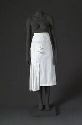 Rok in wit katoen met in de taille een sluiting met knopen en knoopsgaten waarover gespen en linte...